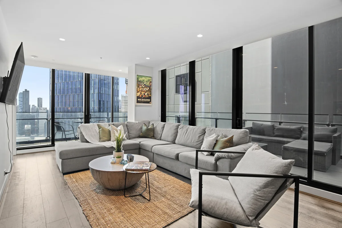A furnished high-rise living room with a large grey sectional sofa, a round timber coffee table and floor-to-ceiling windows over city towers.