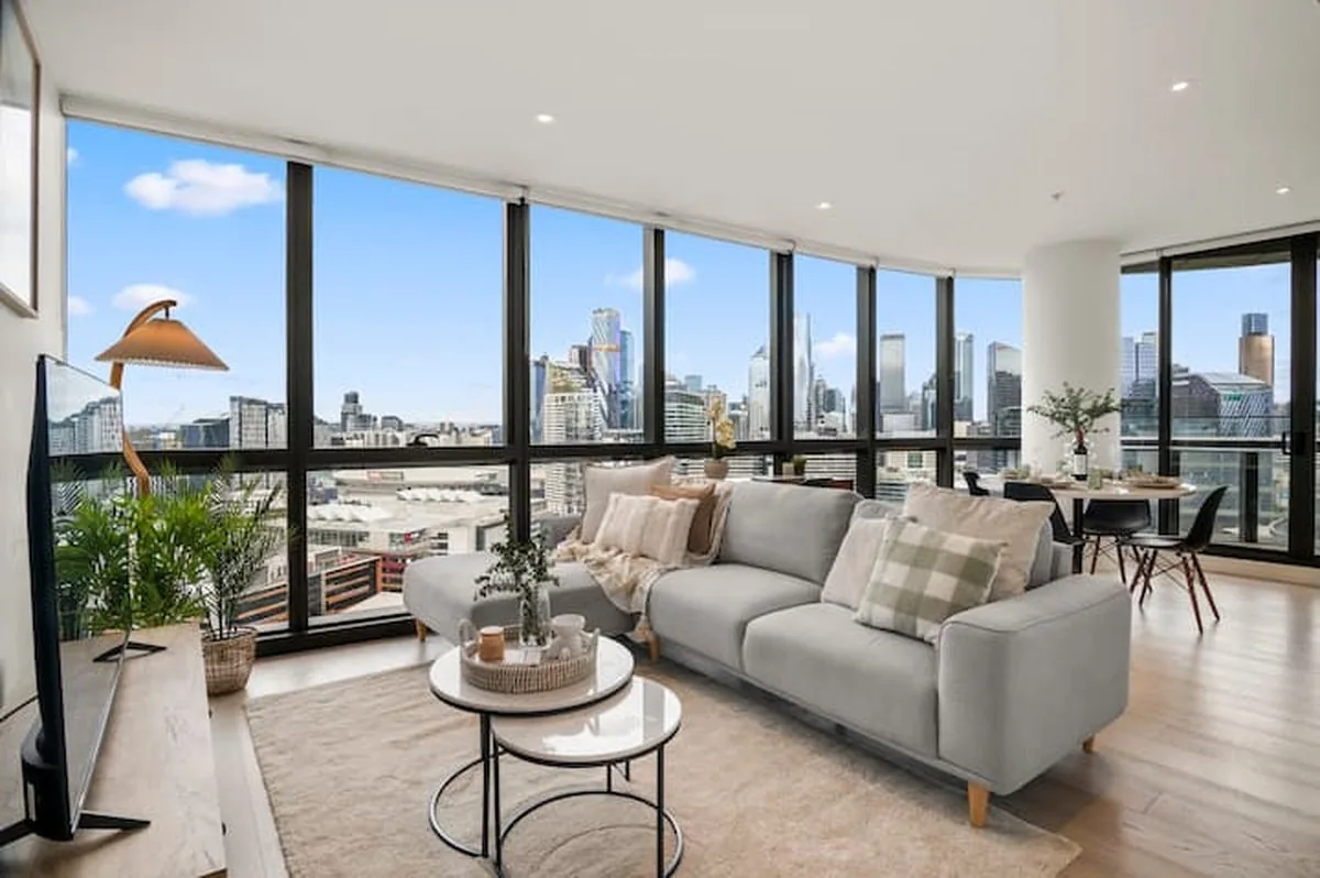 A corner apartment living room with curved floor-to-ceiling windows, a grey sofa and the Melbourne skyline outside.